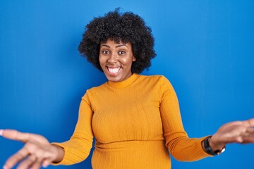 Fototapeta na wymiar Black woman with curly hair standing over blue background smiling cheerful offering hands giving assistance and acceptance.