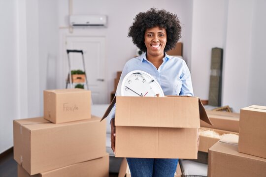 Black woman with curly hair moving to a new home holding cardboard box smiling with a happy and cool smile on face. showing teeth.