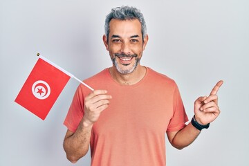 Handsome middle age man with grey hair holding tunisia flag smiling happy pointing with hand and finger to the side