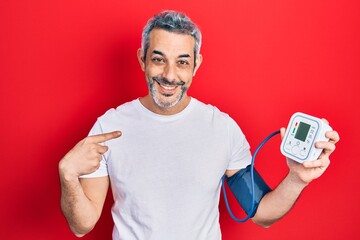 Handsome middle age man with grey hair using blood pressure monitor pointing finger to one self...