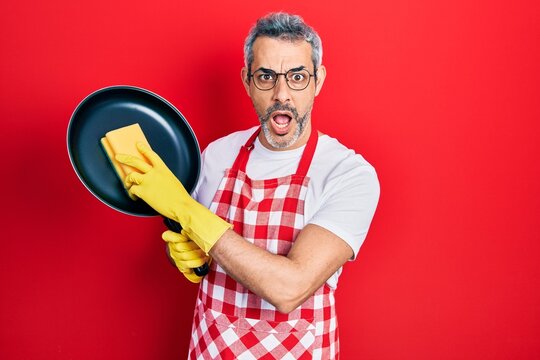 Handsome middle age man with grey hair wearing apron holding scourer washing pan afraid and shocked with surprise and amazed expression, fear and excited face.
