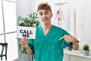 Young caucasian physio man holding call me banner at the clinic pointing finger to one self smiling...