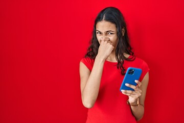 Young brazilian woman using smartphone over red background smelling something stinky and...