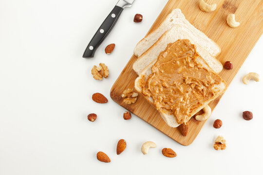 Square bread for toast with peanut butter on a wooden board. Nuts, a knife and a wooden cutting board with a sandwich and slices of bread on a white table.