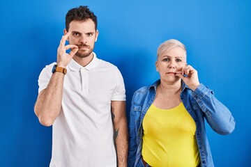 Young brazilian mother and son standing over blue background mouth and lips shut as zip with...