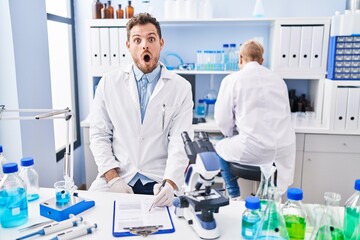 Hispanic man and woman working at scientist laboratory scared and amazed with open mouth for surprise, disbelief face