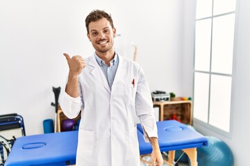 Handsome young man working at pain recovery clinic smiling with happy face looking and pointing to the side with thumb up.