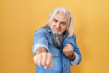 Middle age man with grey hair standing over yellow background punching fist to fight, aggressive...