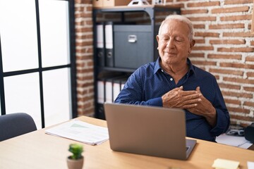 Senior man with grey hair working using computer laptop at the office smiling with hands on chest...