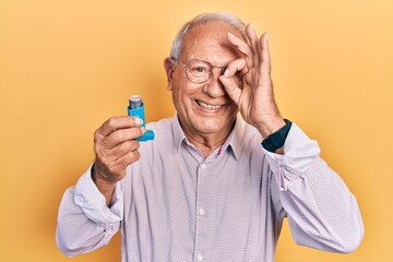 Senior man with grey hair holding medical asthma inhaler smiling happy doing ok sign with hand on eye looking through fingers - obrazy, fototapety, plakaty