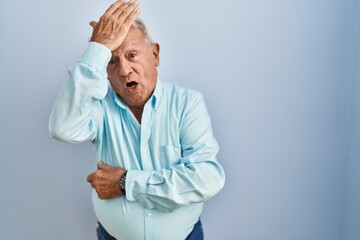 Senior man with grey hair standing over blue background surprised with hand on head for mistake,...