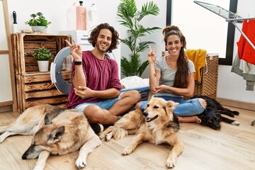 Young hispanic couple doing laundry with dogs smiling with happy face winking at the camera doing...