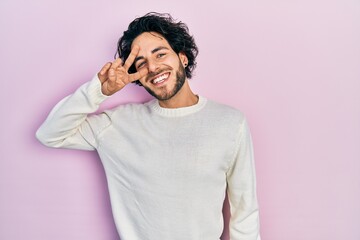 Handsome hispanic man wearing casual white sweater doing peace symbol with fingers over face, smiling cheerful showing victory