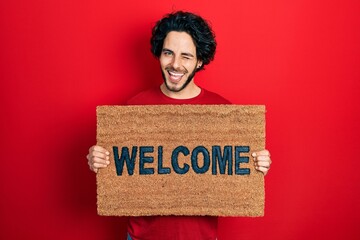 Handsome hispanic man holding welcome doormat winking looking at the camera with sexy expression,...