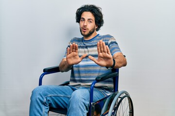 Handsome hispanic man sitting on wheelchair moving away hands palms showing refusal and denial with afraid and disgusting expression. stop and forbidden.