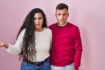Young hispanic couple standing over pink background in shock face, looking skeptical and sarcastic,...
