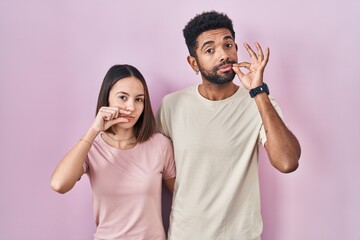 Young hispanic couple together over pink background mouth and lips shut as zip with fingers. secret...