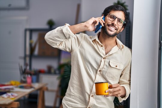 Young hispanic man artist talking on the smartphone drinking coffee at art studio
