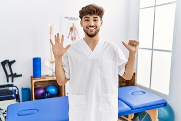 Young arab man working at pain recovery clinic showing and pointing up with fingers number six while smiling confident and happy.