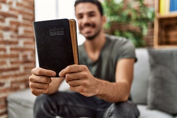 Young hispanic man smiling confident holding bible at home