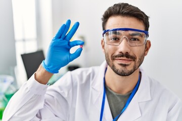 Young hispanic man wearing scientist uniform holding pill at laboratory