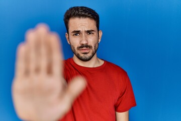 Young hispanic man with beard wearing red t shirt over blue background doing stop sing with palm of the hand. warning expression with negative and serious gesture on the face.