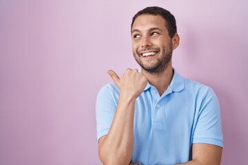 Hispanic man standing over pink background smiling with happy face looking and pointing to the side...