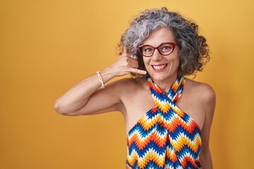 Middle age woman with grey hair standing over yellow background smiling doing phone gesture with hand and fingers like talking on the telephone. communicating concepts.