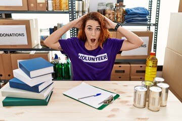 Young redhead woman wearing volunteer t shirt at donations stand crazy and scared with hands on...
