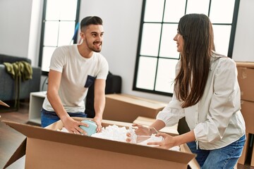 Obraz premium Young hispanic couple smiling happy unpacking cardboard box at new home.