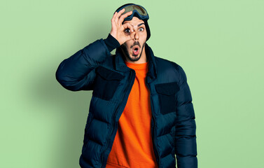 Young hispanic man with beard wearing snow wear and sky glasses doing ok gesture shocked with surprised face, eye looking through fingers. unbelieving expression.