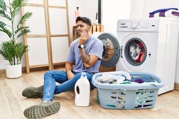 Young hispanic man putting dirty laundry into washing machine touching mouth with hand with painful expression because of toothache or dental illness on teeth. dentist