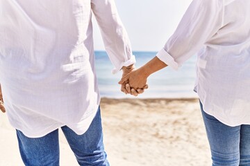 Close up of middle age hispanic couple of husband and wife together holding hands by the beach on a sunny day. Bonding in love on vacation to the seaside.