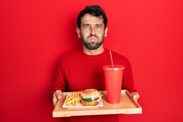 Handsome man with beard eating a tasty classic burger with fries and soda puffing cheeks with funny face. mouth inflated with air, catching air.
