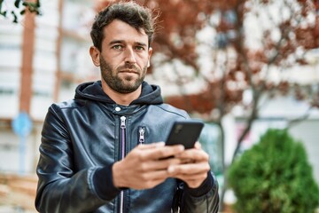Handsome hispanic man with beard with serious face outdoors using smartphone