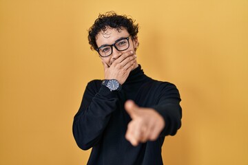Hispanic man standing over yellow background laughing at you, pointing finger to the camera with hand over mouth, shame expression
