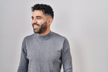 Hispanic man with beard standing over white background looking away to side with smile on face,...