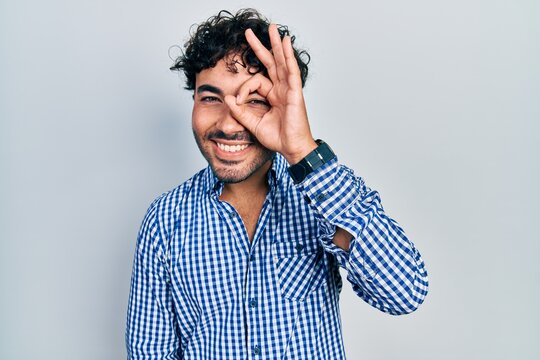 Young hispanic man wearing casual clothes doing ok gesture with hand smiling, eye looking through fingers with happy face.