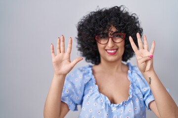 Young brunette woman with curly hair wearing glasses over isolated background showing and pointing up with fingers number nine while smiling confident and happy.