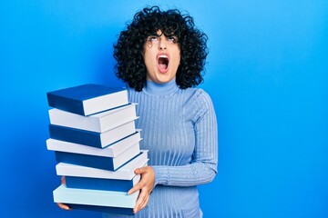 Young middle east woman holding a pile of books angry and mad screaming frustrated and furious,...