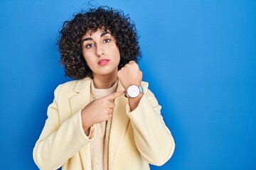 Young brunette woman with curly hair standing over blue background in hurry pointing to watch time,...