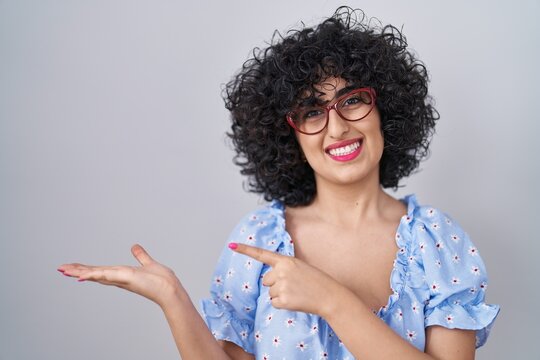 Young brunette woman with curly hair wearing glasses over isolated background amazed and smiling to the camera while presenting with hand and pointing with finger.