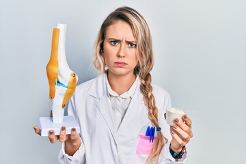 Beautiful young blonde doctor woman holding anatomical model of knee joint and protein powder...