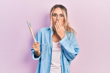 Beautiful young blonde woman holding paintbrushes covering mouth with hand, shocked and afraid for...