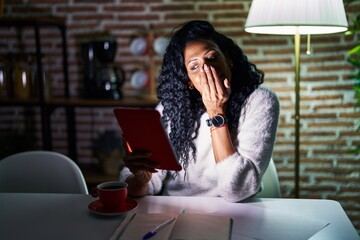 Middle age hispanic woman using touchpad sitting on the table at night laughing and embarrassed...