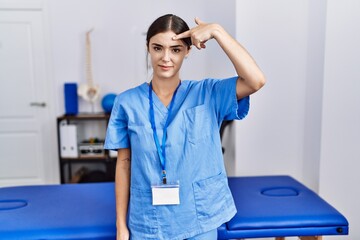 Young hispanic woman wearing physiotherapist uniform standing at clinic pointing unhappy to pimple...