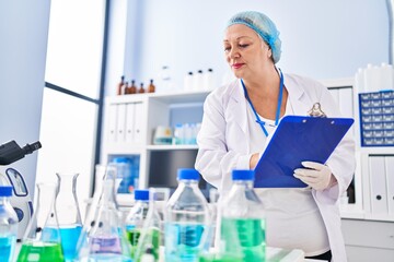 Middle age blonde woman wearing scientist uniform writing on clipboard at laboratory