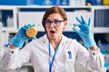 Middle age hispanic woman working at scientist laboratory making vitamin in shock face, looking...
