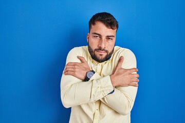 Handsome hispanic man standing over blue background shaking and freezing for winter cold with sad...