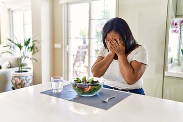 Young hispanic woman eating healthy salad at home with sad expression covering face with hands...